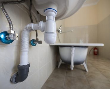 Bottom view of new white washbasin sink connected to sewer on background of bathroom light beige ceramic tiles. Professional plumbing and installation concept.