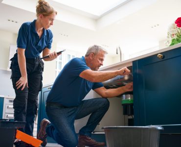 Male Plumber With Female Trainee Using Digital Tablet Fixing Waste Disposal Unit In Kitchen Sink