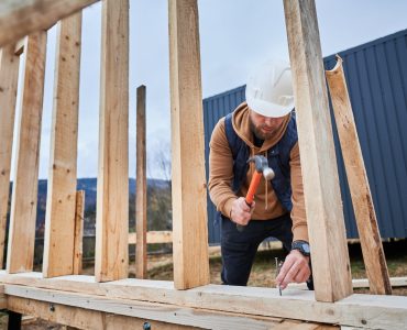 Man worker building wooden frame house on pile foundation. Carpenter hammering nail into wooden plank, using hammer. Carpentry concept.