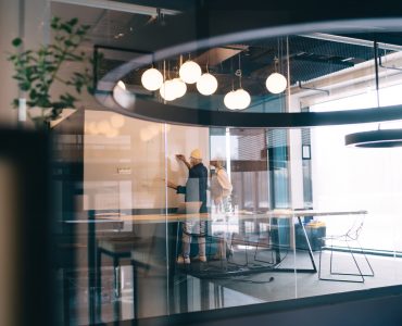 Through glass view of aged male writing on whiteboard while standing with aged female in meeting room with blurred glass wall with reflections of outside images and daylight