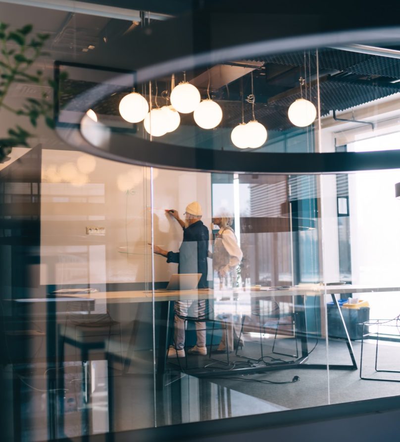 Through glass view of aged male writing on whiteboard while standing with aged female in meeting room with blurred glass wall with reflections of outside images and daylight