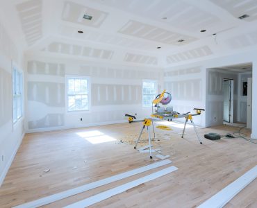Spacious, unfinished room with drywall partially installed miter saw set up in center on stand, surrounded by wood trim pieces construction debris at hardwood floor.