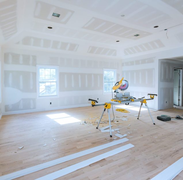Spacious, unfinished room with drywall partially installed miter saw set up in center on stand, surrounded by wood trim pieces construction debris at hardwood floor.