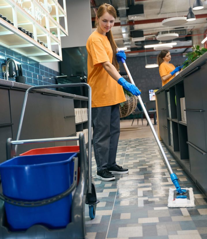 Young woman cleaning the floor with a mop, her colleague is in the background