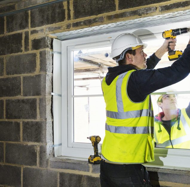 A workman on a construction site, a builder in hard hat using an electric drill on a window sill.